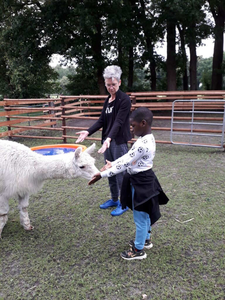 Ein Kind das neben einer Therapeutin steht und die einem Lama die Hand hin streckt.